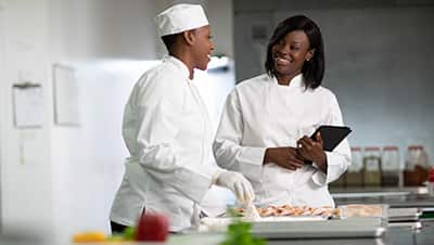 A leader holding a tablet and speaking with one of her employees who is preparing food in a restaurant's kitchen.