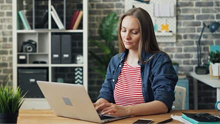 A woman sitting at a table typing on a laptop in an office setting.