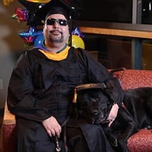 Ricardo Scarello wearing his cap and gown with his service dog, Puck, laying beside him.
