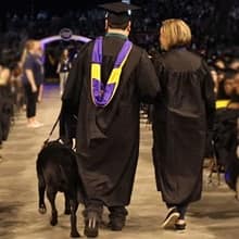 Ricardo Scarello walking down an aisle at SNHU commencement ceremony with his service dog, Puck, and a woman assisting him.