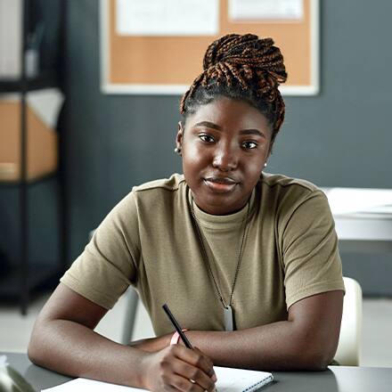 A young woman with braided hair sitting at a desk while holding a pen with a notebook in front of her and a bulletin board visible in the background.