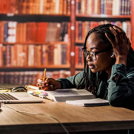 A student sitting at a desk in a library taking notes from a laptop