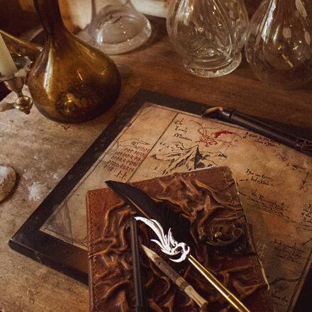 A quill and notebook sitting atop a map on weathered desk, surrounded by antique looking objects.