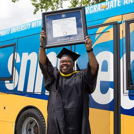 Clad in graduation regalia and standing in front of an SNHU bus, Lorenzo Mateo '19, holds up a framed diploma after earning a BS in Information Technology.