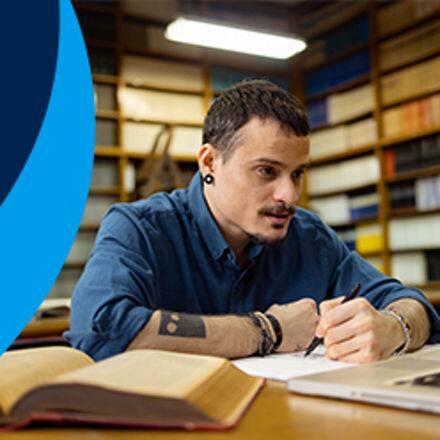 A student sitting in a library working on their english degree with a stack of books to the left on a blue background.