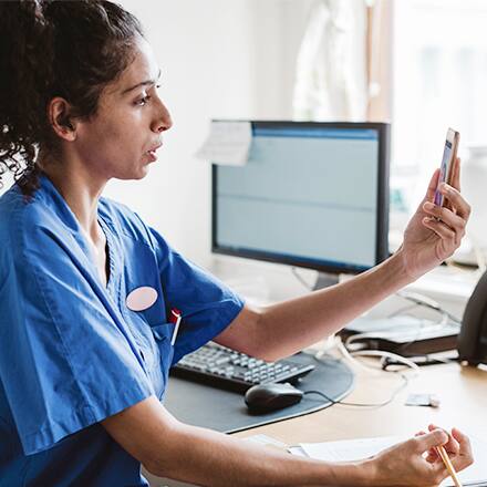 A woman with dark hair in a blue medical shirt looking at a phone and working in the role of telehealth in nursing