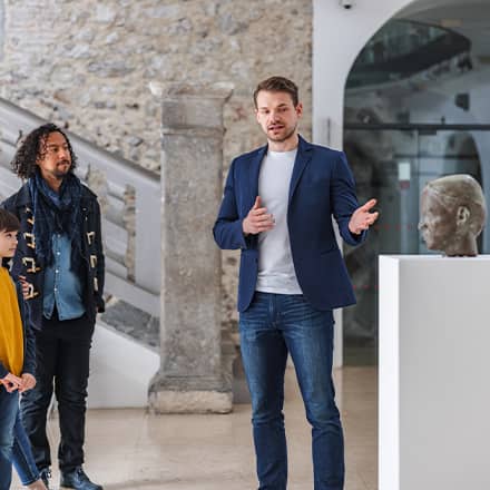 A curator with a master's in history, gesturing to a sculpture of a head during a museum tour.
