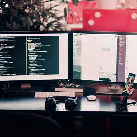 The desk of a web developer with two desktops open on development work along with a pair of headphones and a phone.