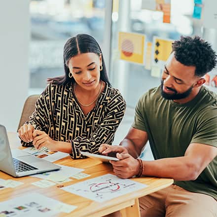 Two brand managers sitting together and looking at a tablet with printed documents and a laptop open in front of them.