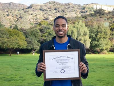 Keyon Tuiteleleapaga, who earned a bachelor's and master's degree at SNHU, standing in a field holding his framed diploma.