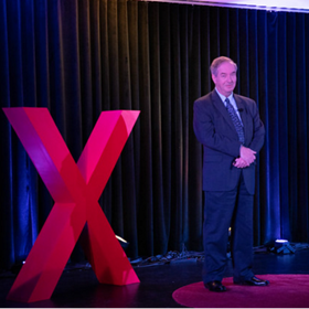 Anthony Knopps speaking at TEDxSNHU in 2024, to the right of a large, red letter X