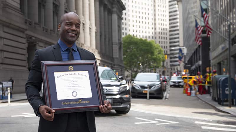 Jean Besson, who earned a BS in business degree online, holding his 2014 SNHU diploma in a city setting.