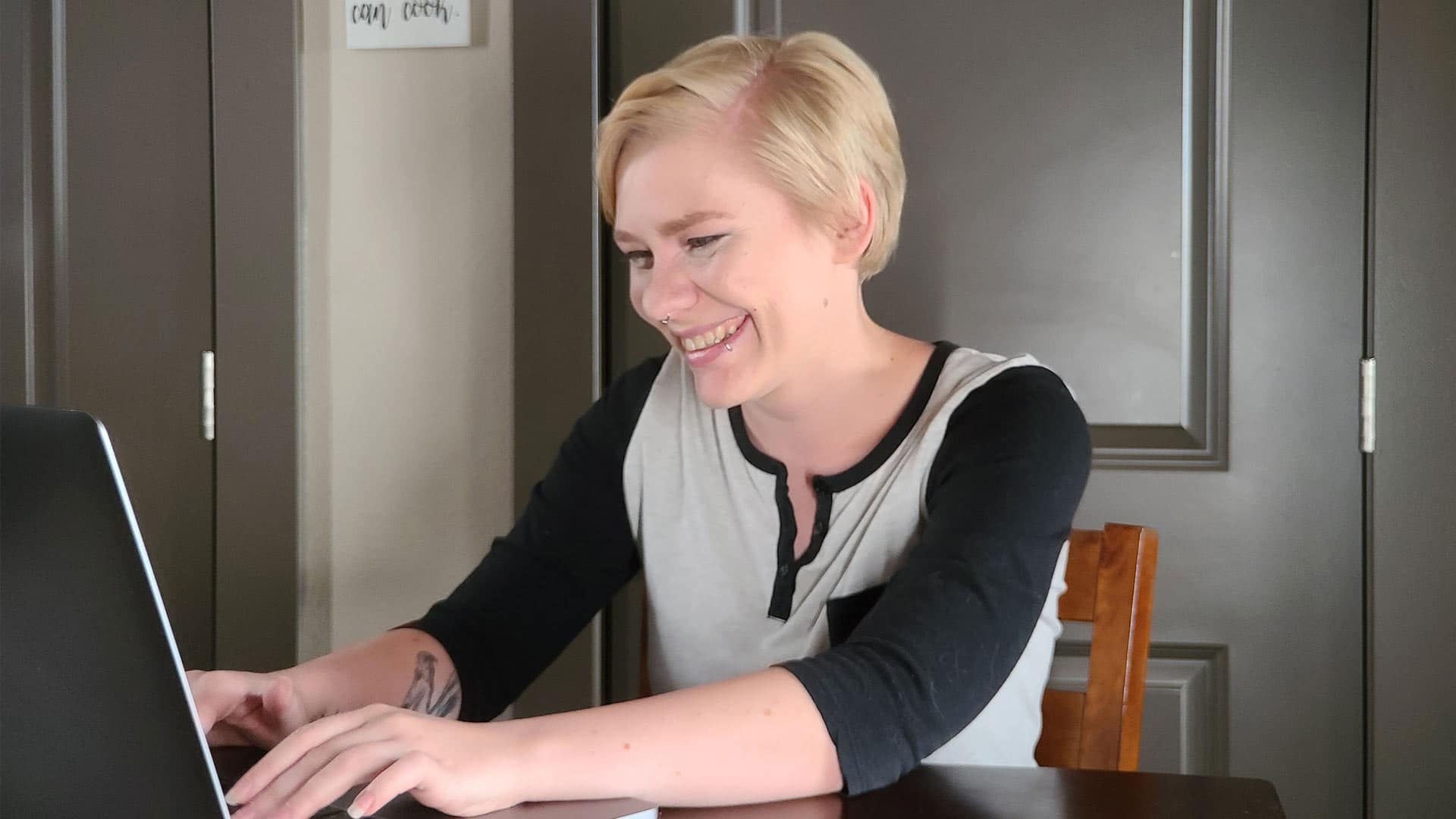 Jolene Stetz, who earned a marketing degree in 2021, wearing a grey and black long sleeve T-shirt smiling and working on her laptop.