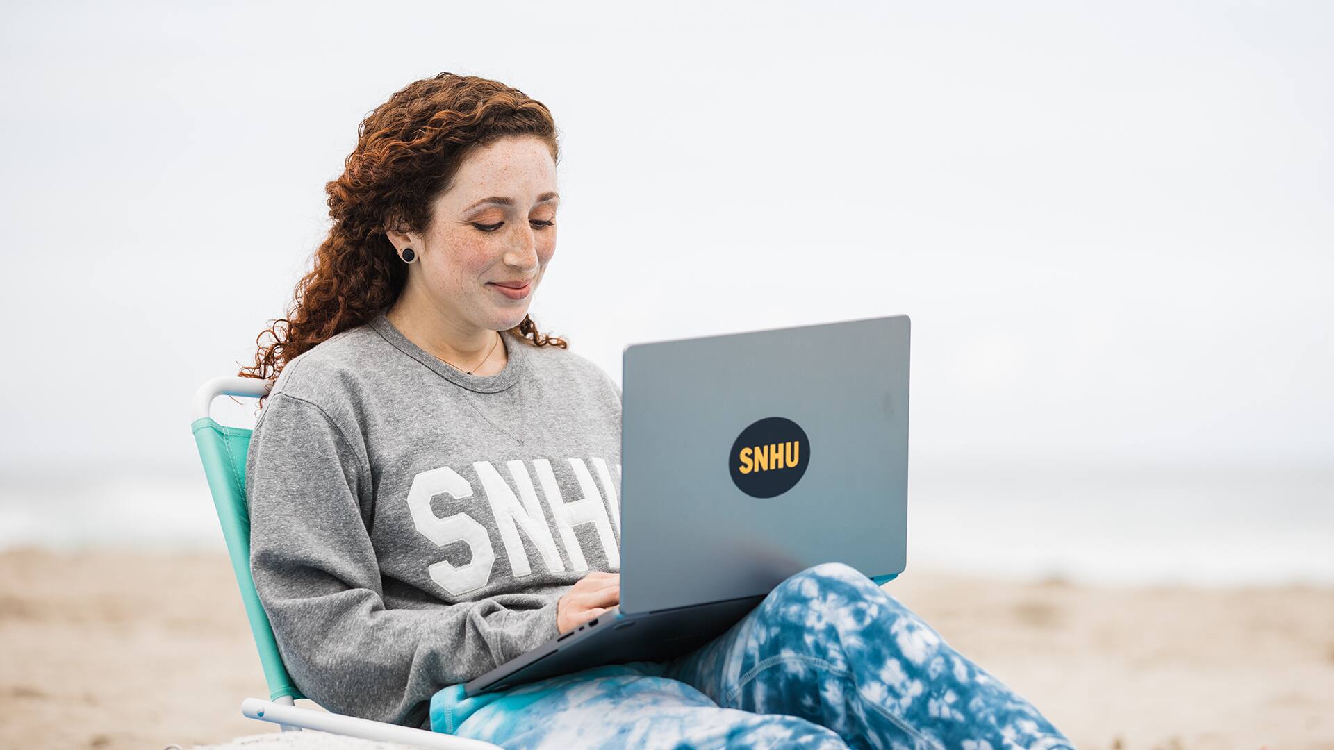 Mariel Embry, who earned her degree in creative writing and English, wearing a grey SNHU sweatshirt sitting in a foldable chair on the beach and working on her laptop.
