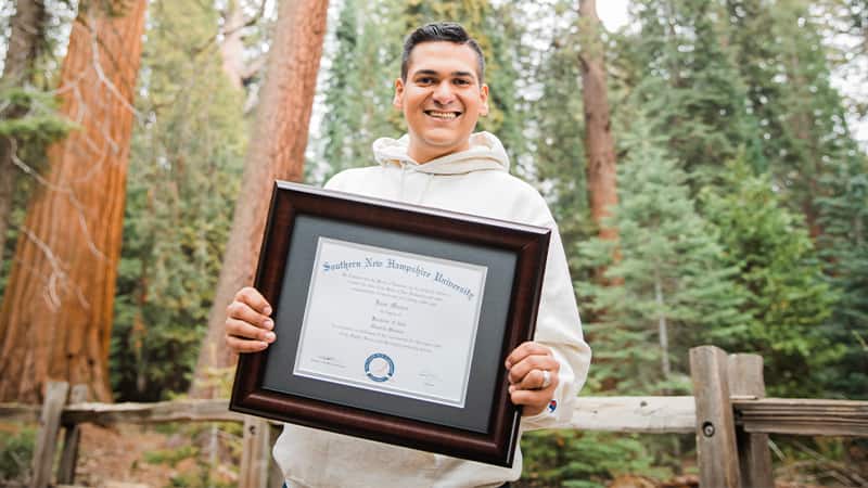 Juan Munoz, who eanredh is BA in General Studies in 2022, smiling and holding his framed SNHU degree in front of a wooden post and rail fence and dark green evergreen trees.