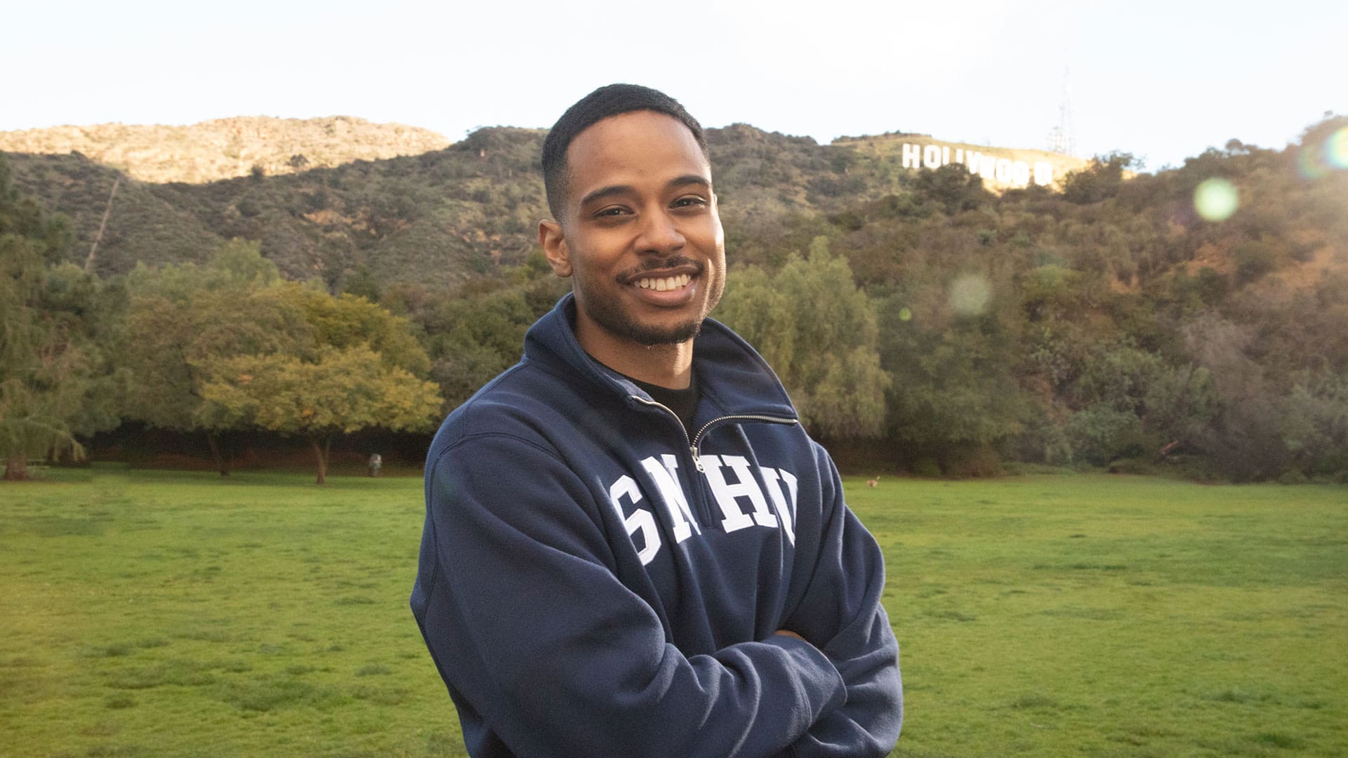 Keyon Tuiteleleapaga, who earned his degree from SNHU in 2024, wearing a dark blue SNHU sweatshirt smiling with his arms folded standing in a grass field with trees and the Hollywood sign in the distance behind him.