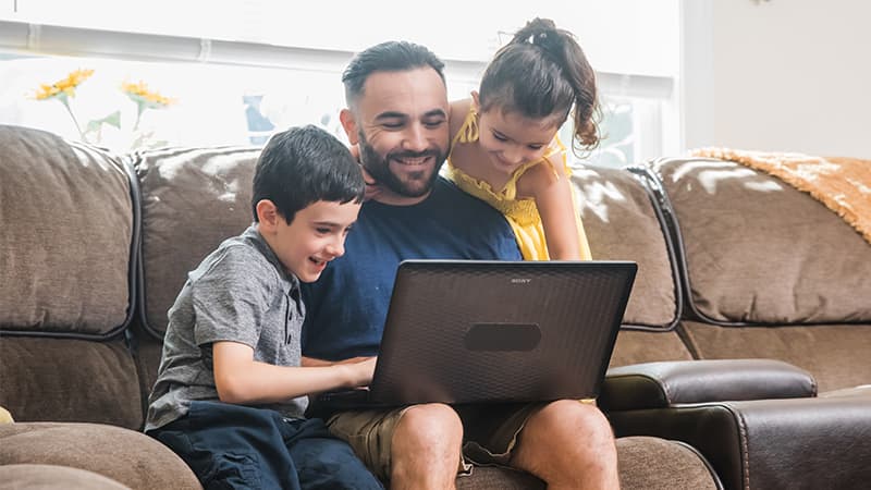 Stephen Goulakos, working from a laptop on his couch while his two school-age children play around him.