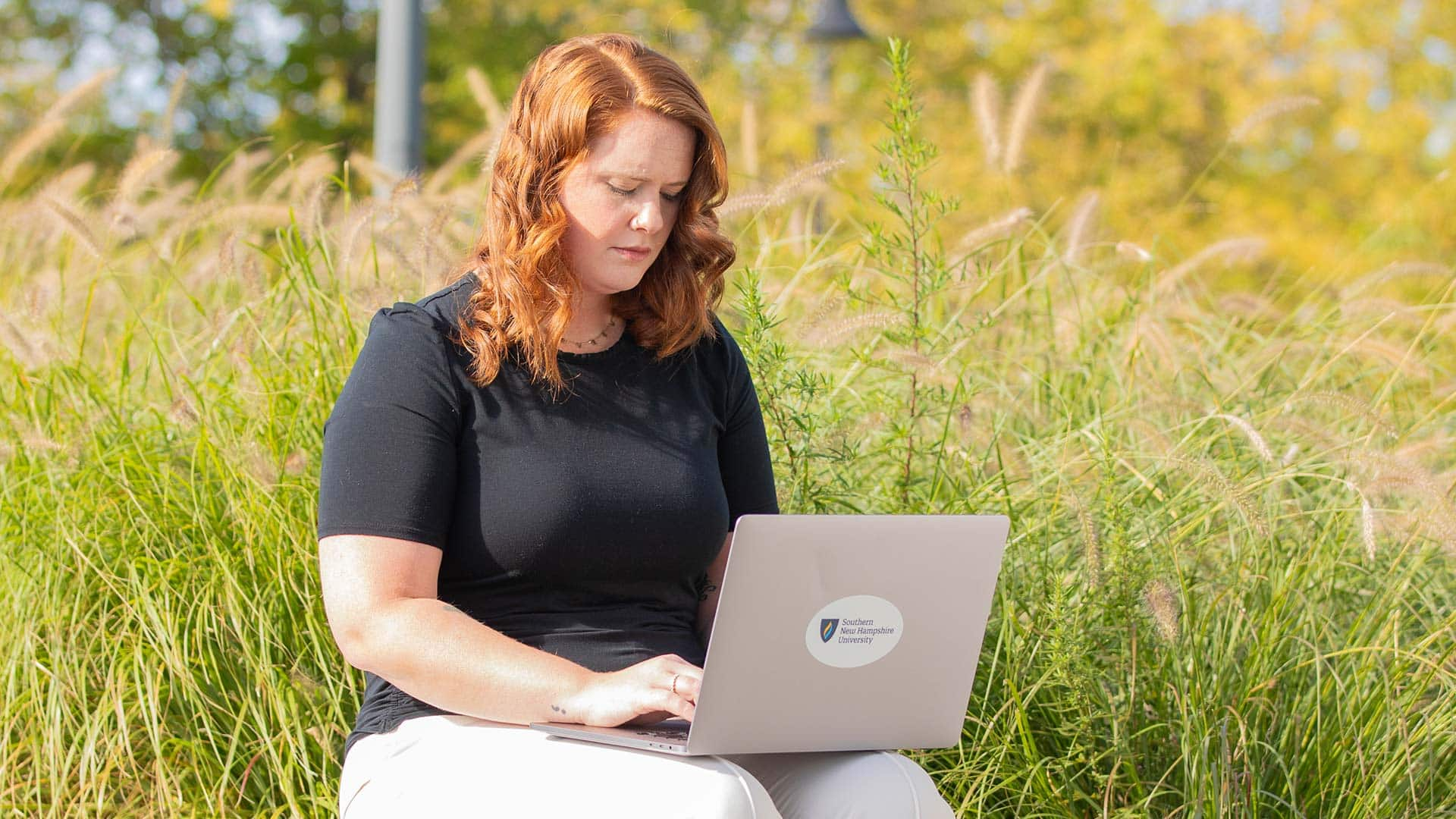 An SNHU student sitting outside in a field while typing on a laptop with the Southern New Hampshire University logo on it