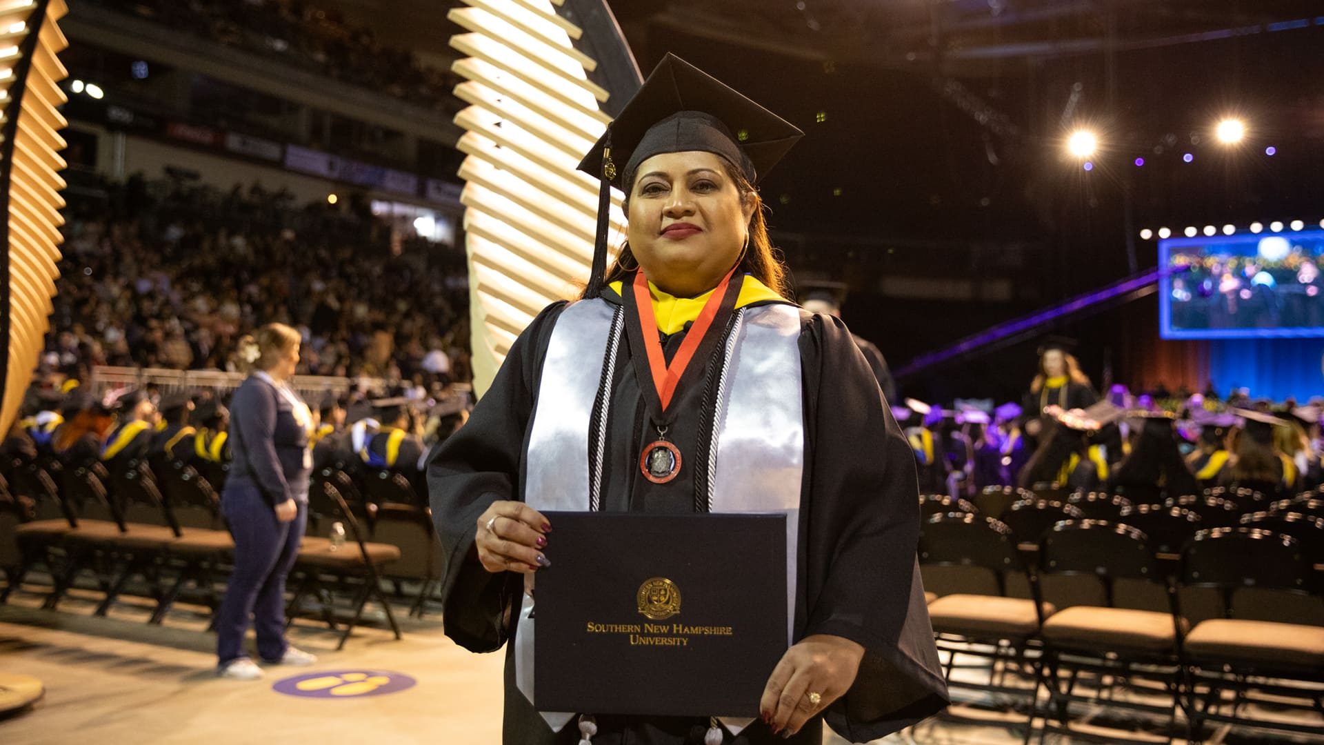 Karie Lamb, who earned her degree in 2023, wearing her cap and gown, holding her SNHU diploma in front of her while standing in the SNHU Arena at a Commencement ceremony.