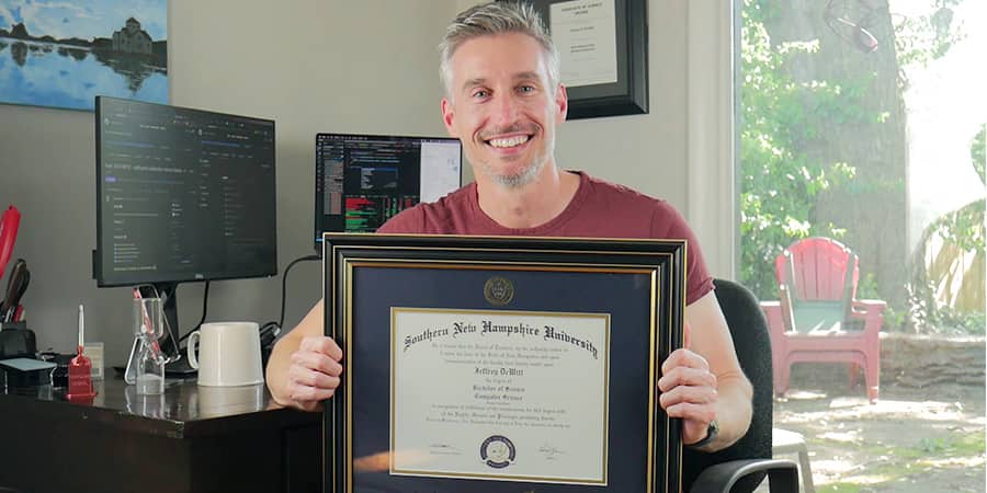 Jeffrey Dewitt, sitting at a work desk holding his Bachelor of Science in Computer Science diploma from Southern New Hampshire University.