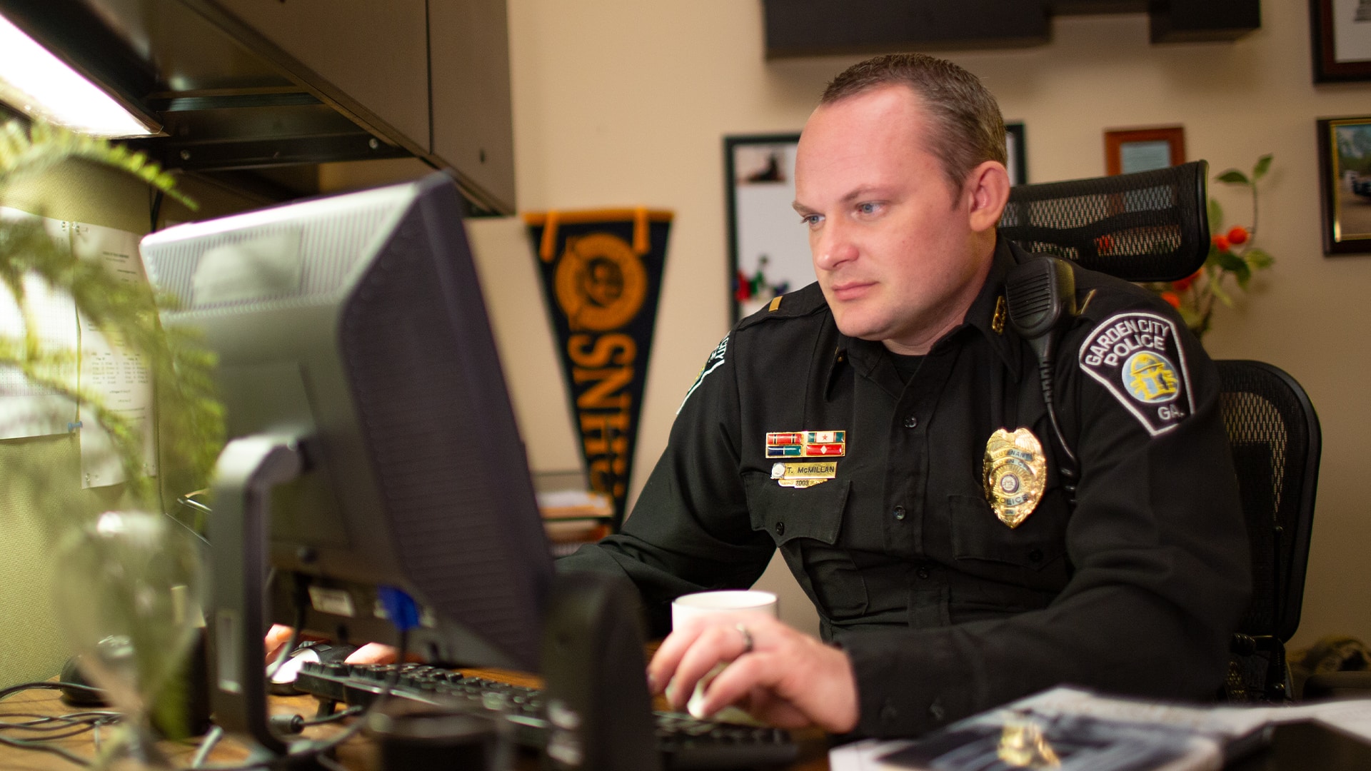 Tim McMillan, who earned his degree from SNHU in 2015, wearing his police uniform in an office working on a computer with an SNHU pennant on the wall in the background.