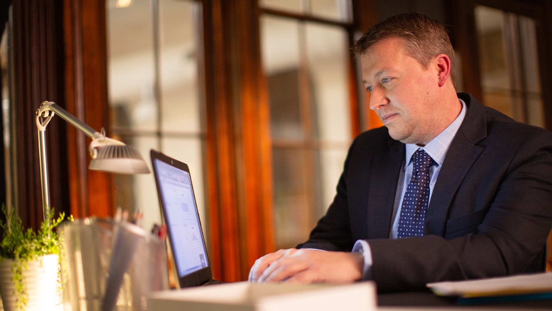 Brock Ehlers, who earned his degree from SNHU in 2014, typing on a laptop with a small desklamp and potted  plant in front of him.