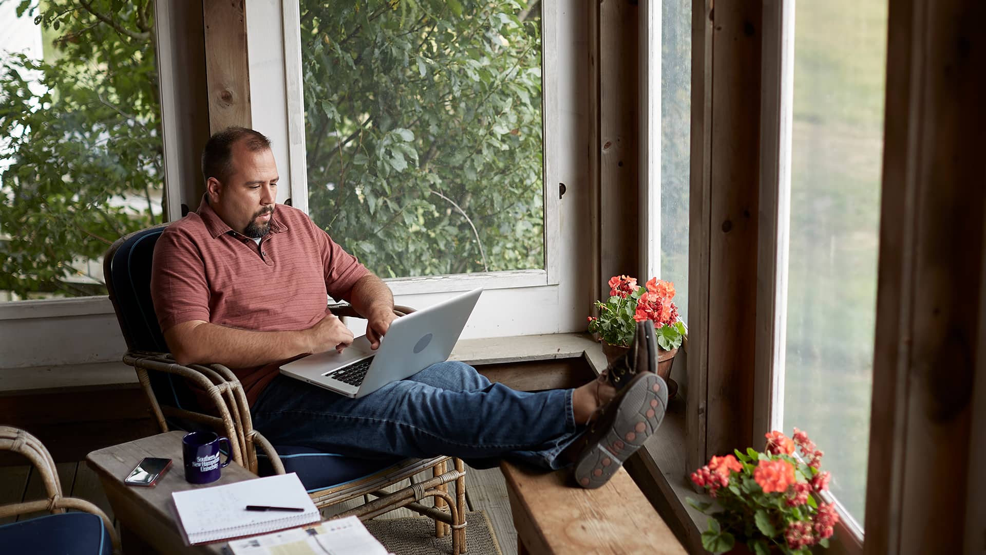 Raymond Gammon, who earned a degree in data analytics in 2020, sitting with his feet up in an indoor porch working on a laptop with an open notebook, smartphone and SNHU coffee mug next to him.