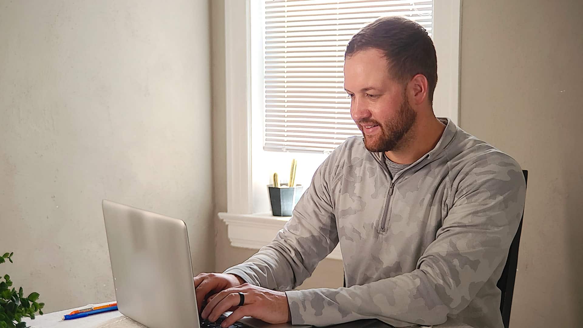 Wyatt Martensen, who earned his degree from SNHU in 2021, sitting at a table working on his computer with a  window covered by a white venitian blind in the background.