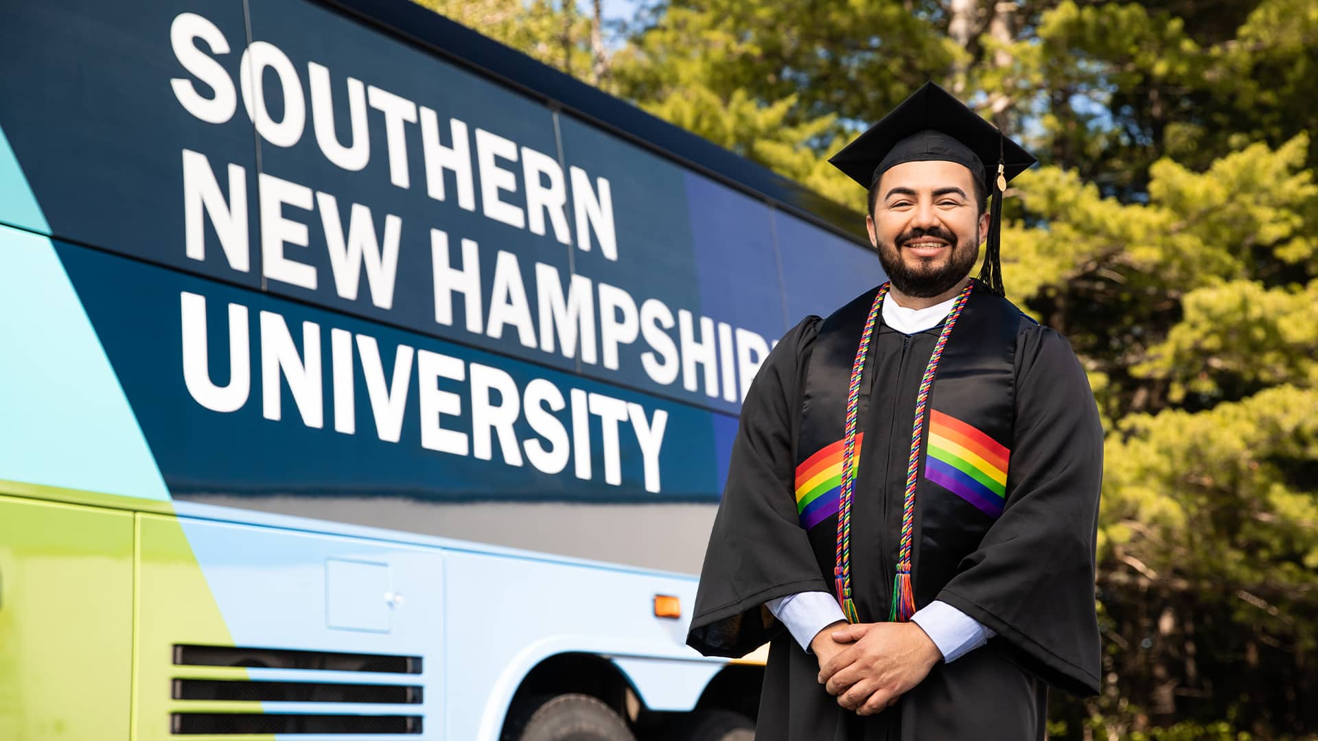 Jesús Súarez, who earned his degree from SNHU in 2021, wearing his cap and gown standing in front of the SNHU-branded bus.