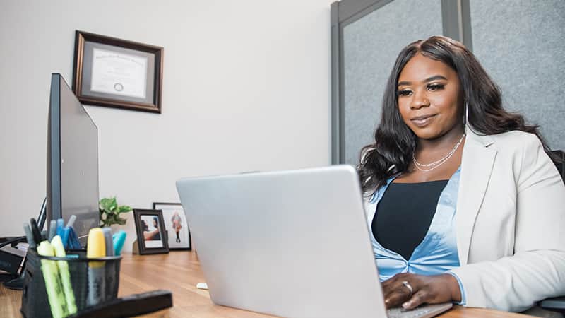 Tanzania Fair, who earned her degree from SNHU in 2020, in an office working on a laptop with her framed  SNHU degree on the wall behind her.
