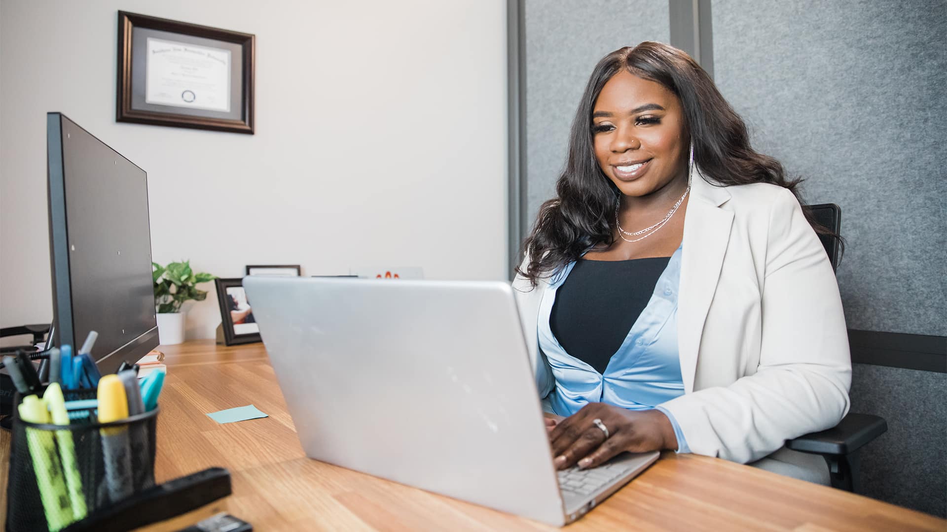 Tanzania Fair, who earned her degree from SNHU in 2020, sitting in an office working on a laptop with her framed diploma on the wall in the background.