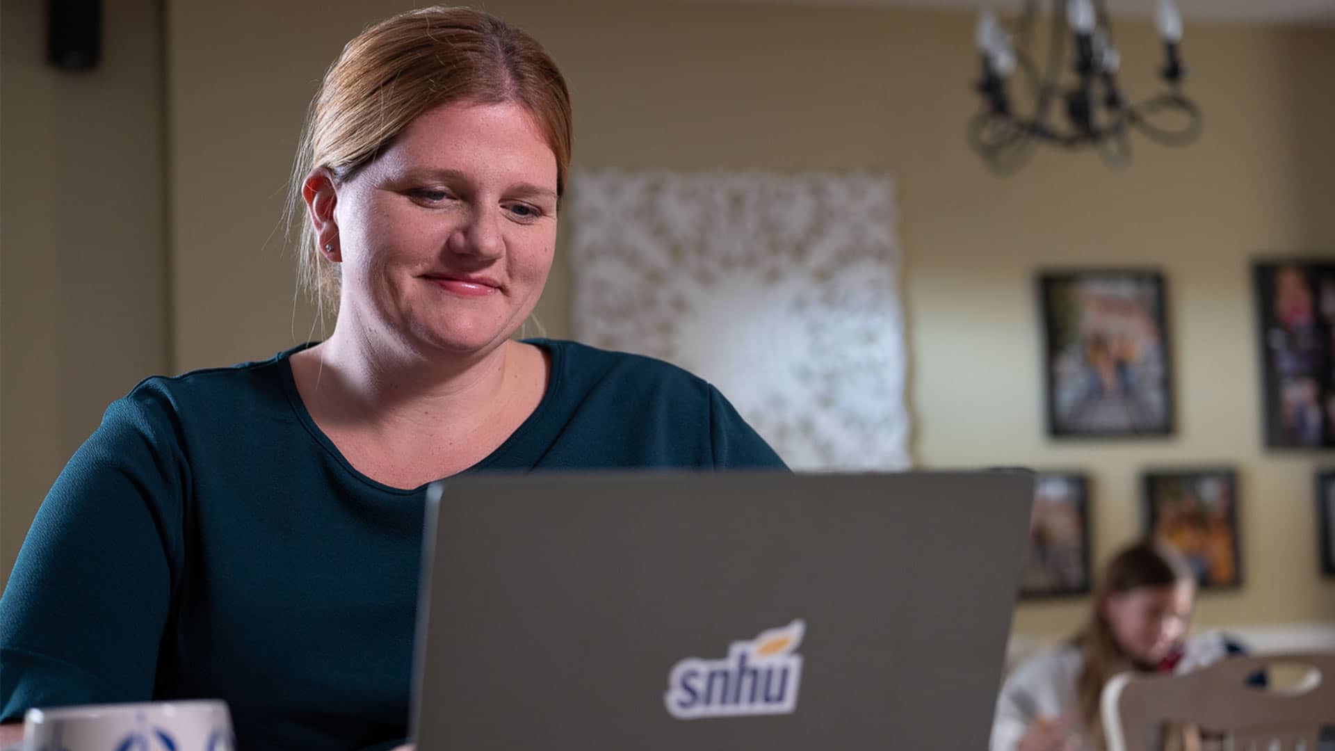 "Shelly Villa, who earned her degree from SNHU in 2019, working on her laptop with an SNHU sticker on the front and  a girl seated in the background."
