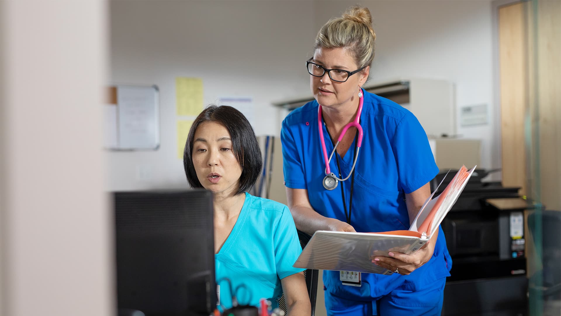 Kristina Libby, who earned her degree from SNHU, wearing blue scrubs and a pink stethoscope, leaning over the shoulder of another woman at a computer.