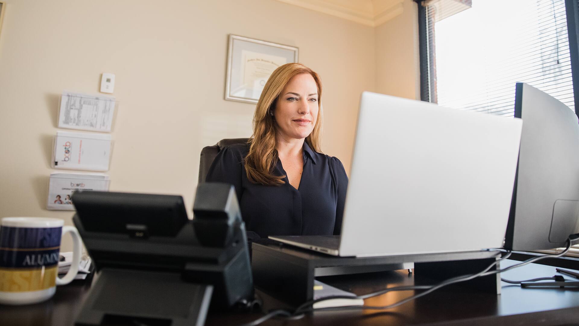 Nikki Bennett, who earned her MBA from SNHU, sitting at a desk in her office looking at her laptop with her framed diploma on the wall in the background.