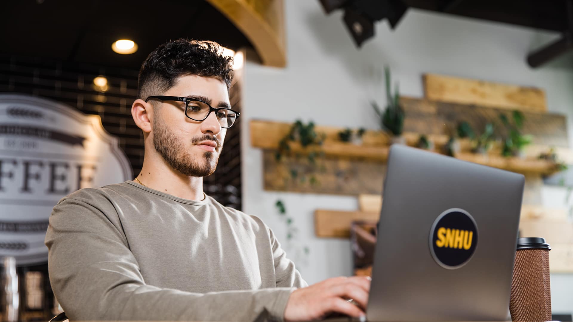 Naeem Jaraysi, an SNHU alum, wearing a grey sweatshirt in a coffee shop working on his laptop with an SNHU sticker on the front.