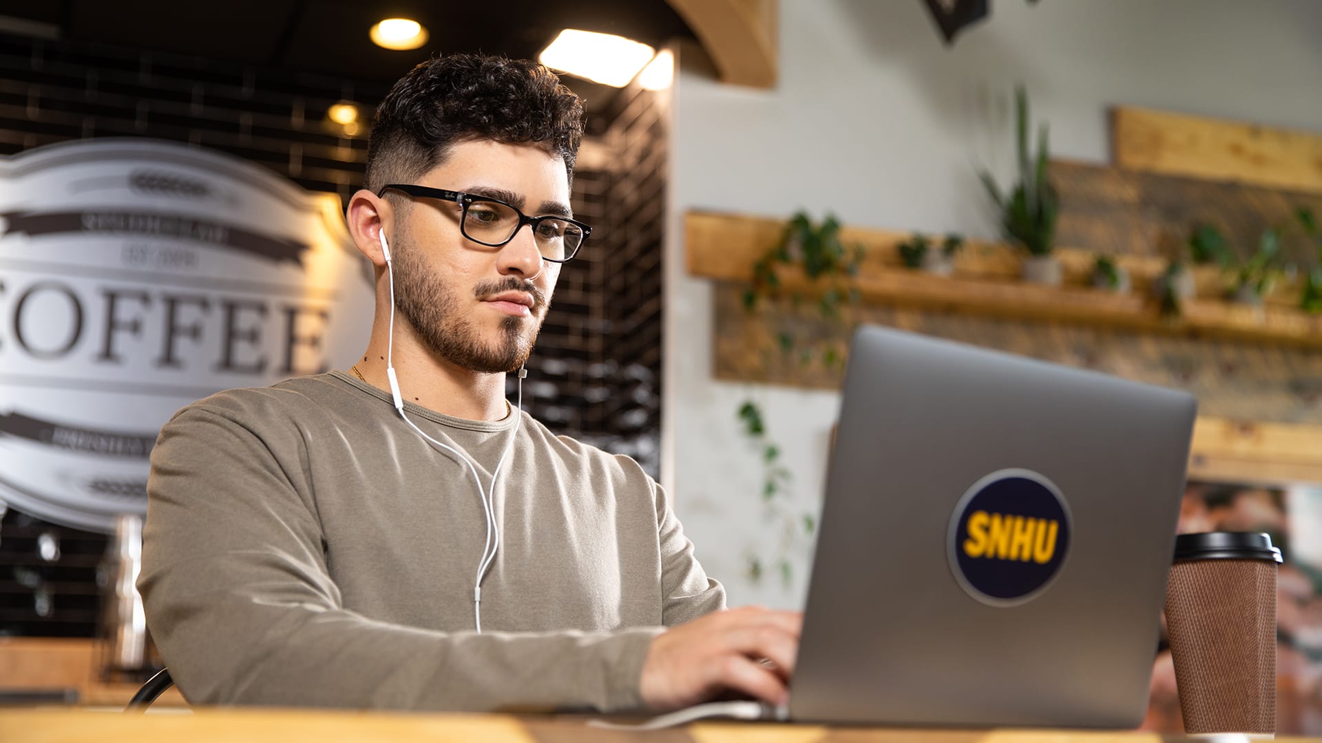 Naeem Jaraysi, who earned his degree from SNHU, wearing ear pods working on his laptop with an SNHU sticker on the front in a coffee shop.