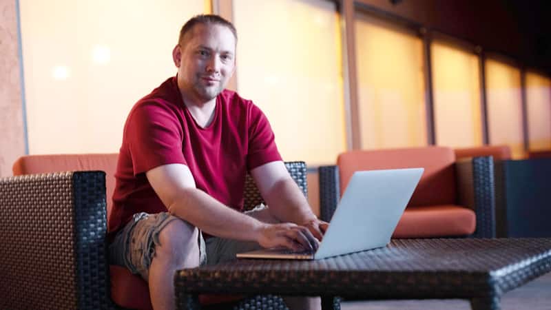 John Roos, who earned his degree from SNHU in 2018, wearing shorts and a red T-shirt, sitting in a chair and  typing on his laptop.