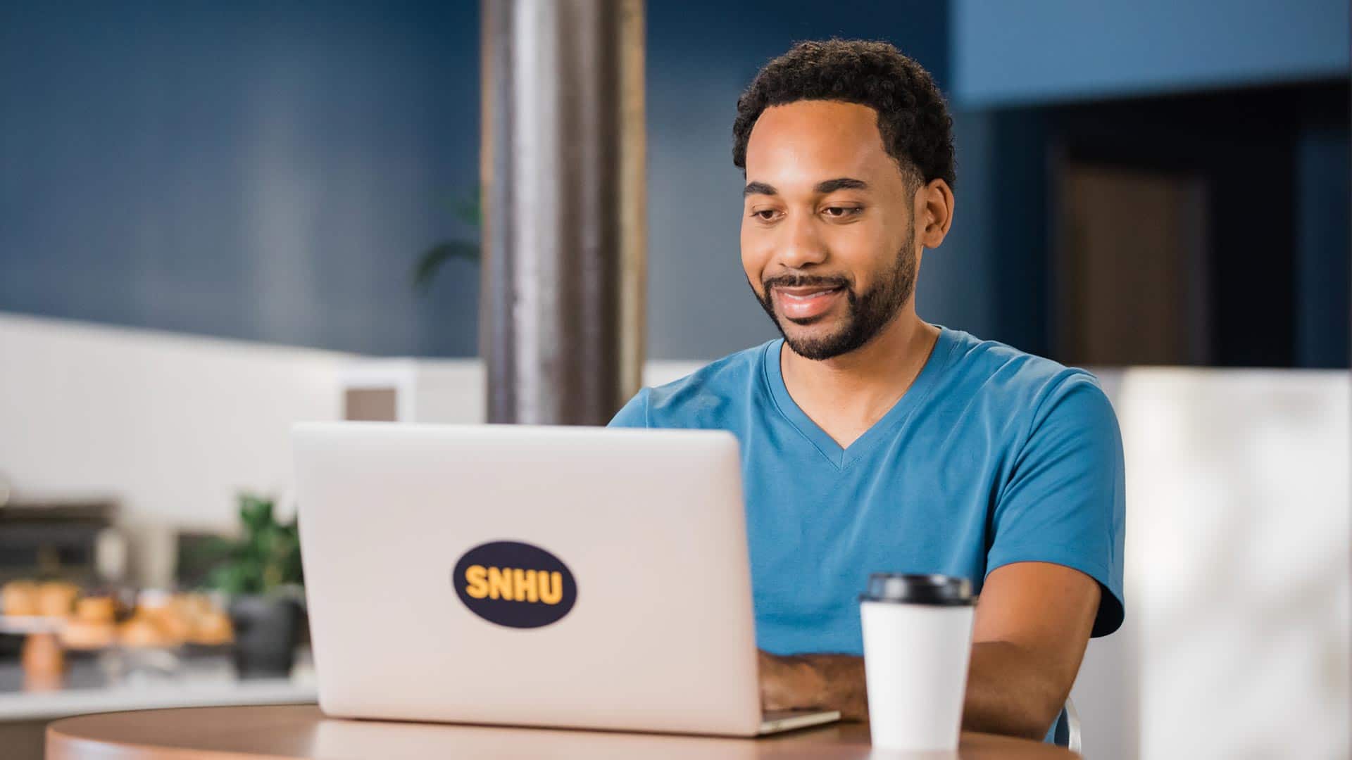 Gary Walker, who earned his degree from SNHU in 2022, sitting in a coffee shop working on his laptop that has an SNHU sticker on the cover.