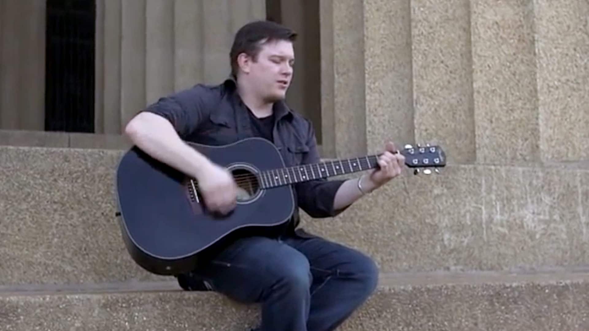 Russ Kettle, who earned his degree from SNHU in 2017, sitting on a set of stone steps playing a black acoustic guitar.