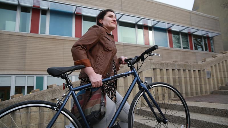 Zepure Kurumlian, who earned her degree from SNHU in 2016, wearing a brown jacket and carrying a bicycle up a stone staircase.