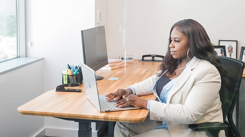 Tanzania Fair, who earned her MS in Accounting in 2020, wearing a light colored pants suit, sitting at a desk in an office working on her computer.