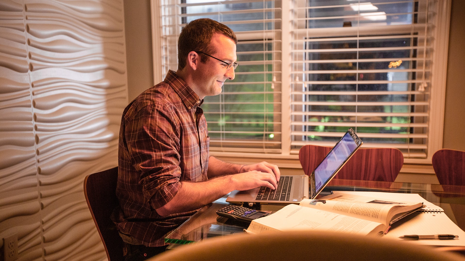 Chris Eldridge, who earned a cybersecurity degree, sitting in a home office working on a laptop with an open notebook, text book and calculator on the desk beside him.