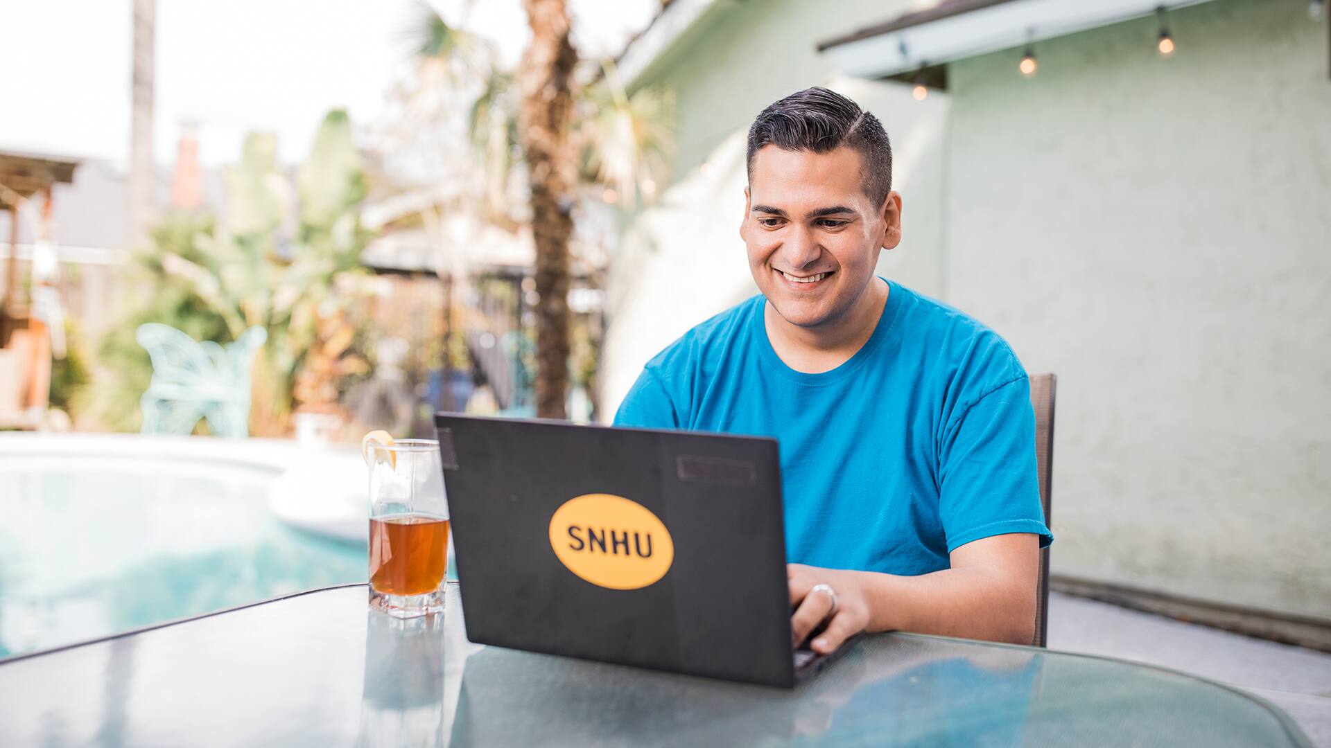 Juan Munoz, who earned his degree from SNHU, sitting at an outdoor table typing on a laptop with an SNHU sticker on the front and a glass of iced tea next to him and a pool in the background.