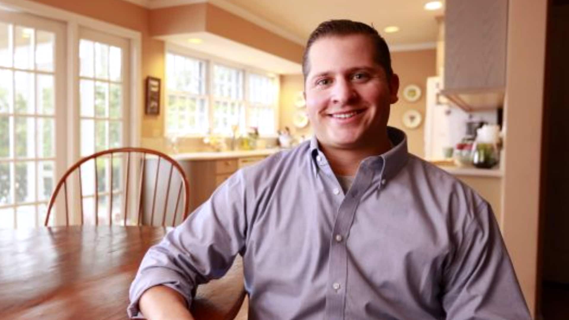 Peter Forcelli, who earned his master's in healthcare administration in 2015, sitting at a kitchen table in a buttondown dress shirt.
