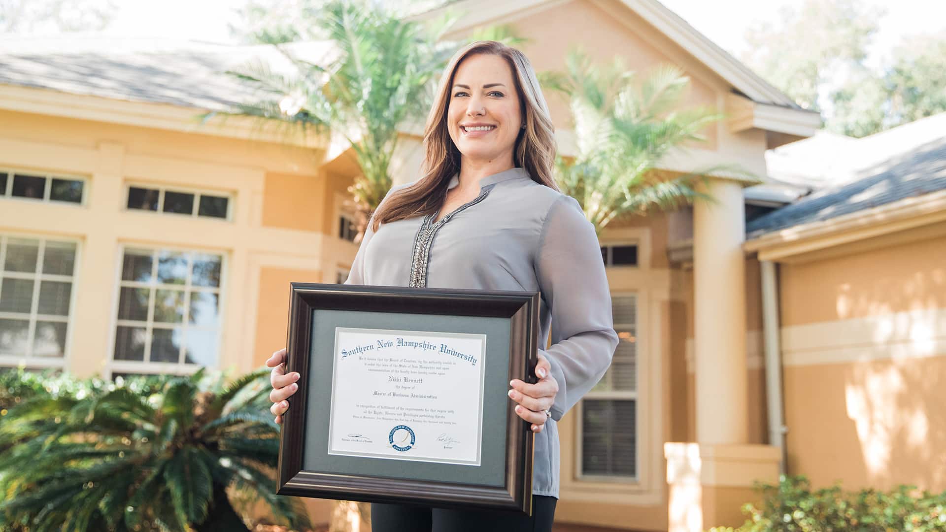 Nikki Bennett, who earned her dgree at SNHU, standing in her front yard holding her framed SNHU diploma