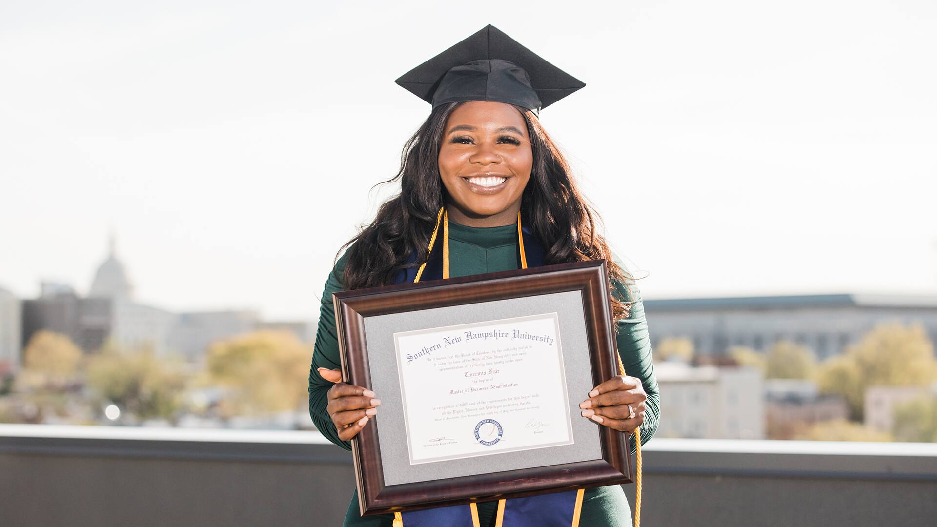 Tanzania Fair, who earned her degree from SNHU in 2020, wearing her cap and gown and holding her framed diploma in front of her.