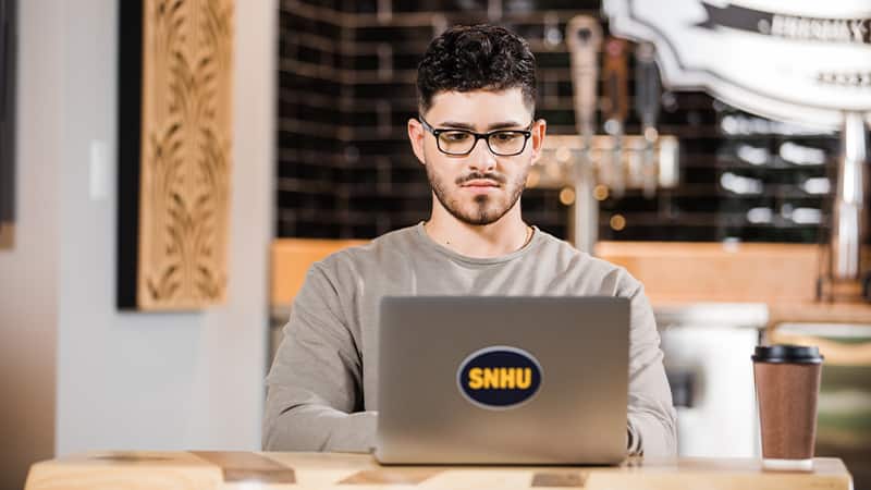 Naeem Jaraysi, who earned his degree from SNHU in 2020, working on his laptop with an SNHU sticker on the cover and a coffee cup on the table next to him.