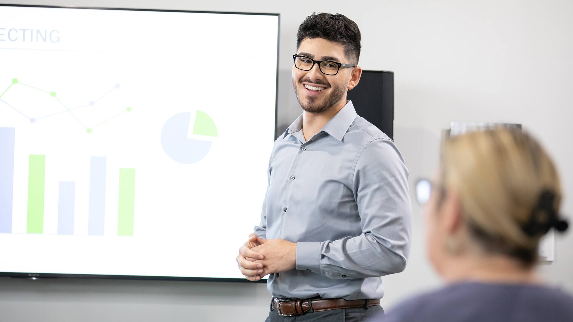 Naeem Jaraysi, who earned his degree from SNHU in 2020, speaking to a woman while standing in front of a projection screen displaying green and blue line, bar and pie charts.