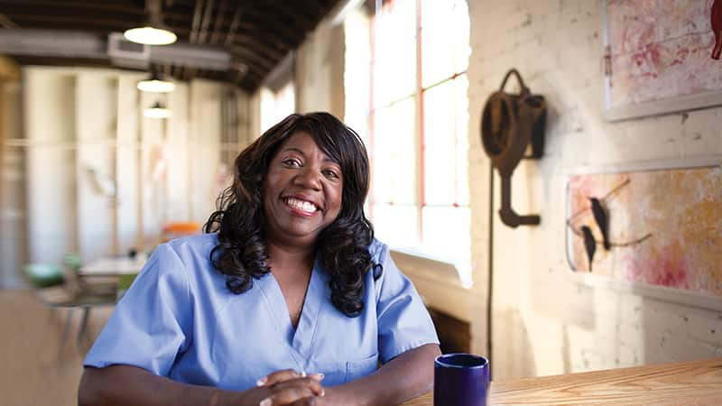 Dana Aulds, who earned her degree from SNHU, wearing blue scrubs and sitting at a small table with a cup of coffee next to her.