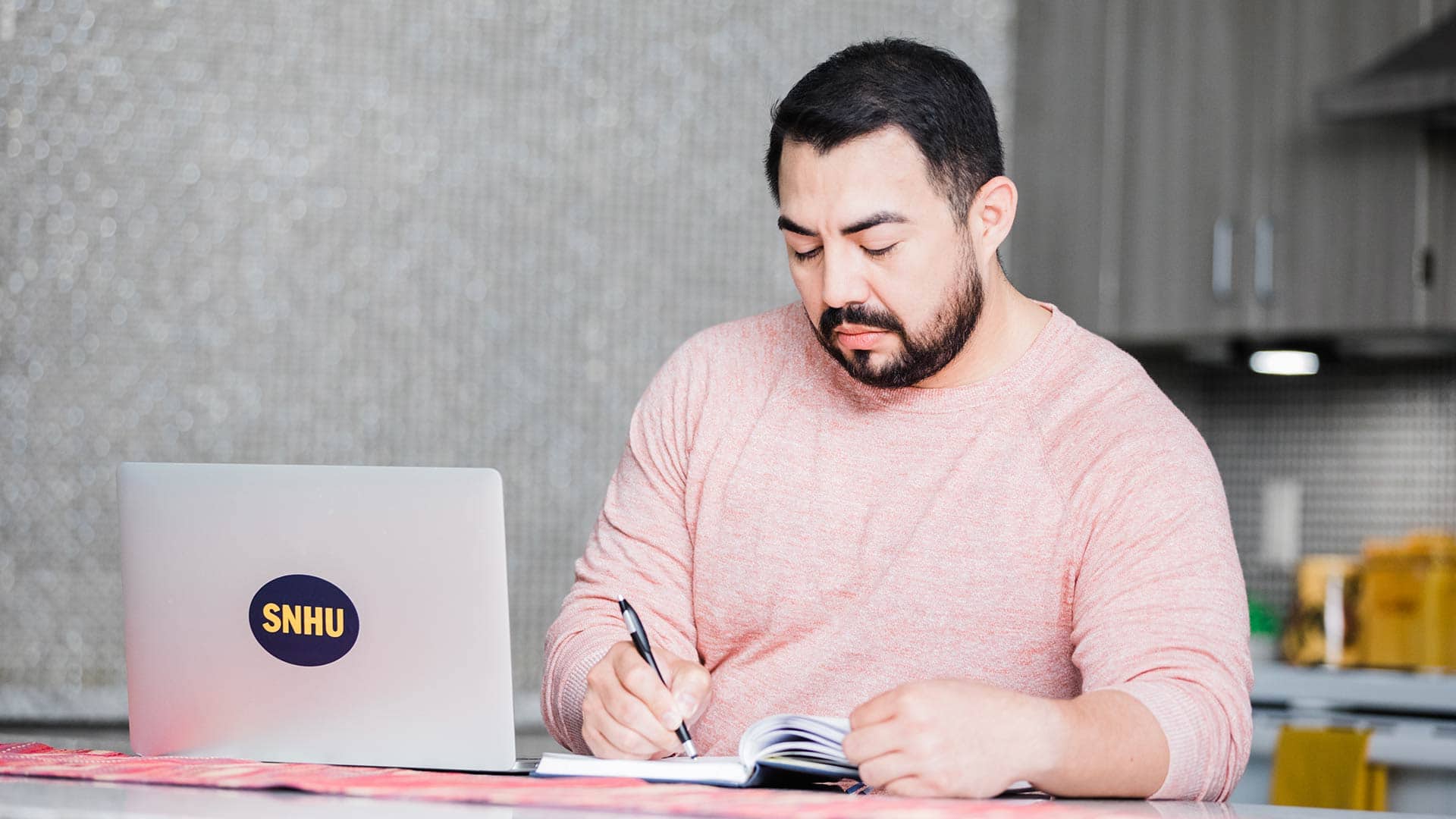 Jesus Suarez, who earned his degree from SNHU in 2021, in his kitchen writing in a small notebook with his open laptop with a SNHU sticker on the front next to him. 